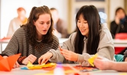Two female students smiling and engaged in making colourful paper crafts at a table